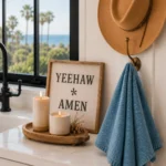 A tan cowboy hat hangs from a brass wall hook beside a blue linen kitchen towel, while a framed “Yeehaw Amen” sign and two candles sit on a wooden tray atop a white quartz countertop near a black-framed window overlooking palm trees and the ocean.