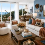 A coastal-inspired living room features a white sofa with rust and blue accent pillows, two cream swivel chairs, a wood coffee table, woven rug, leather chair, ocean-view balcony, and framed horse artwork above a blue accent wall.